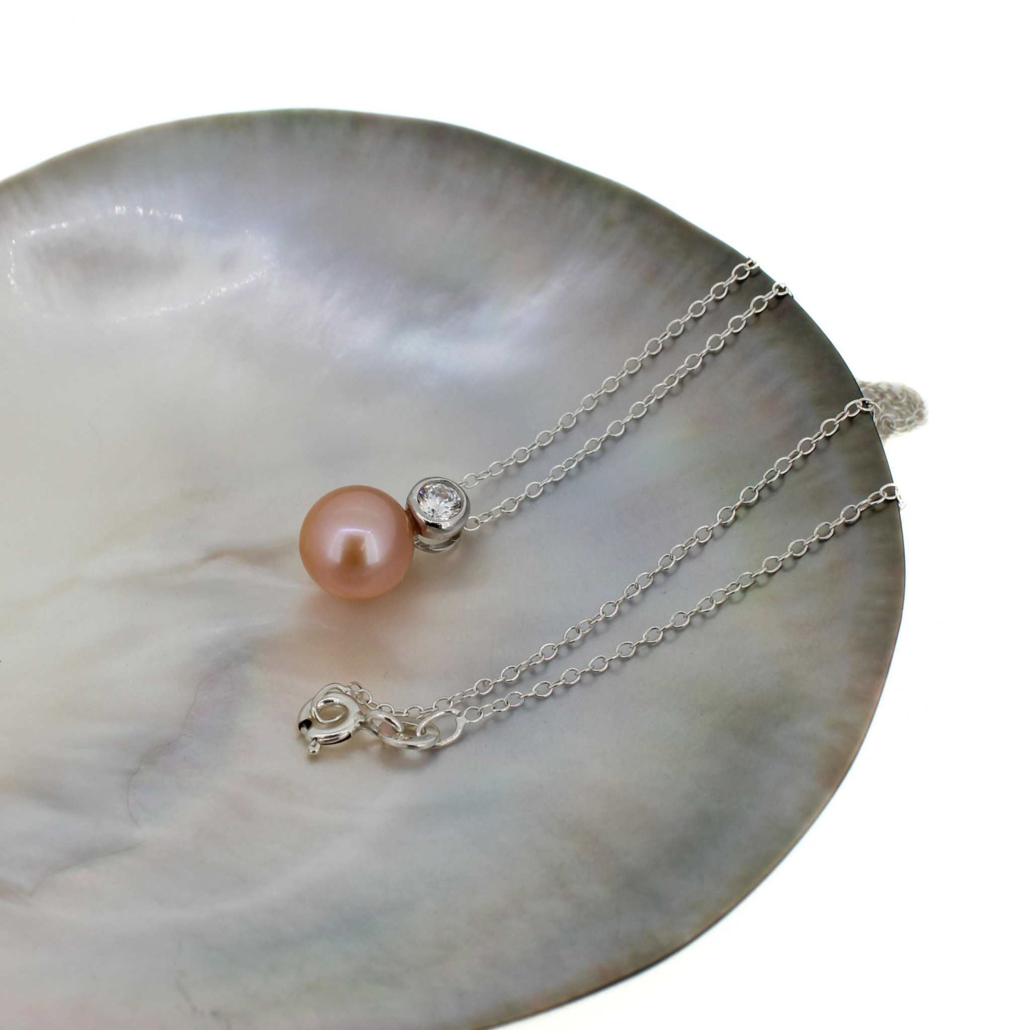 A silver chain necklace with a single peach-pink pearl and a clear bezel-set stone, displayed on a polished iridescent mother-of-pearl dish against a white background.