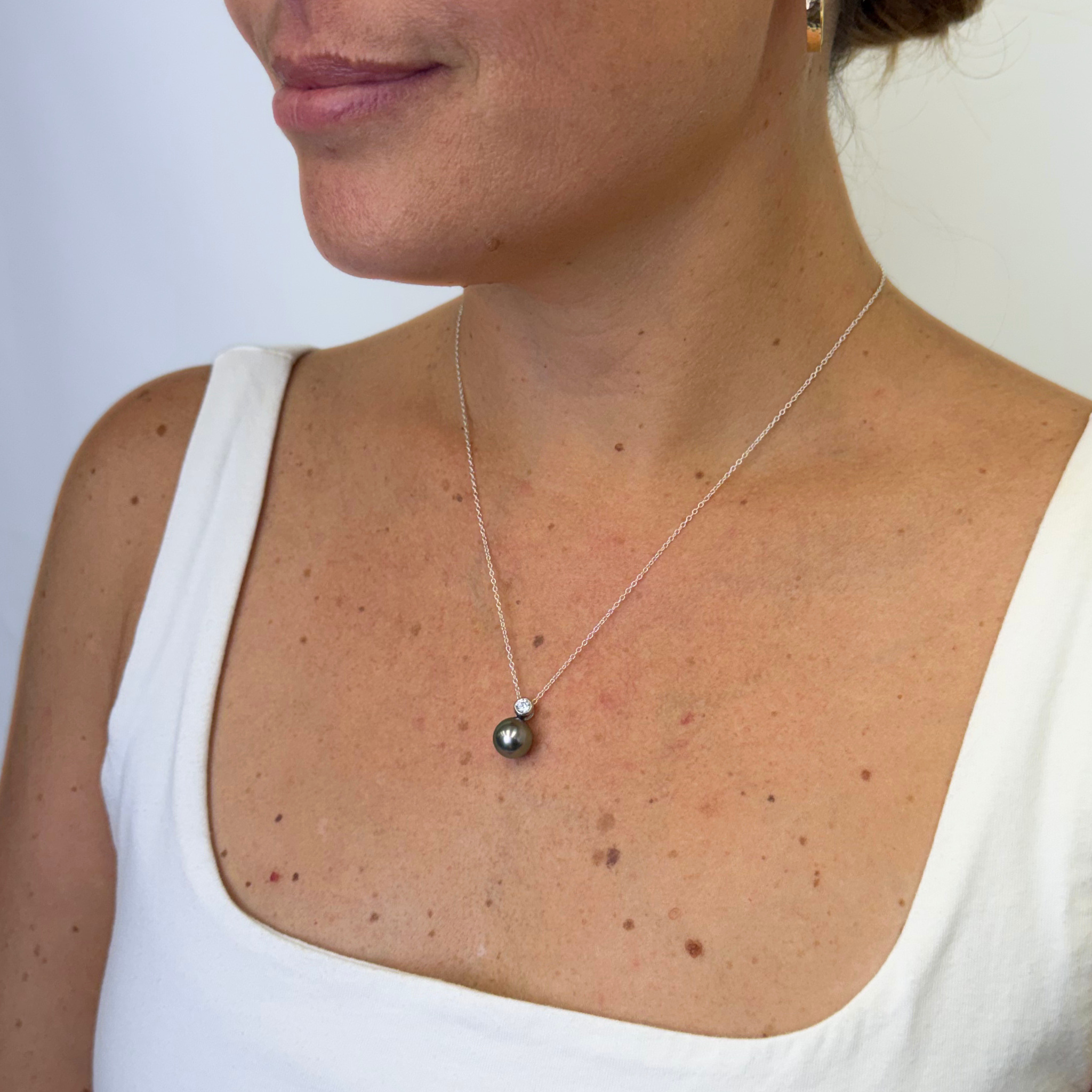 A close-up of a woman wearing a delicate silver chain necklace with a dark, round pearl pendant, paired with a white tank top.