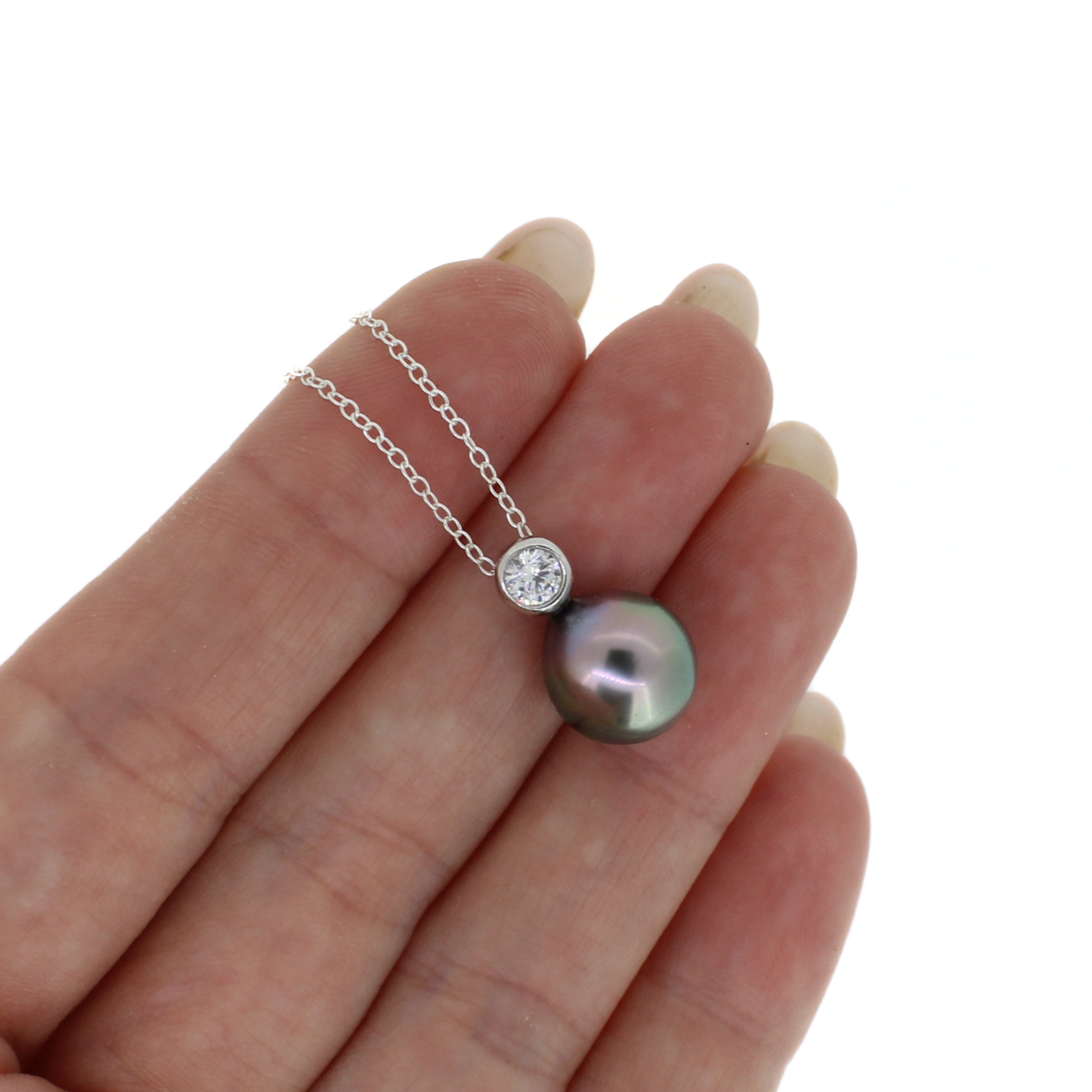 A silver chain necklace with a round iridescent pearl pendant and a small clear bezel-set stone, shown resting on a person's fingers against a white background.