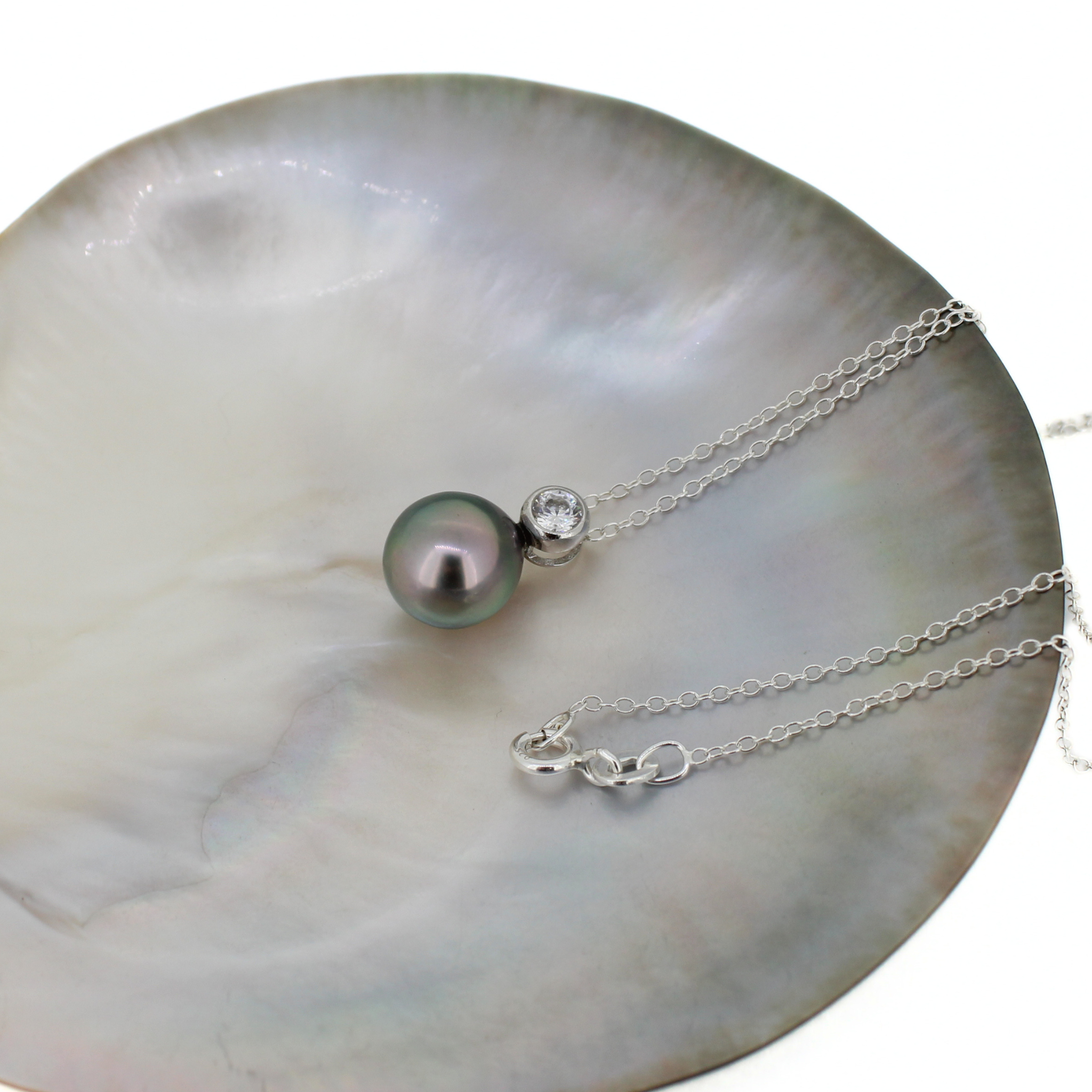 A silver necklace with a dark gray-black Tahitian pearl and a small clear stone accent, displayed on a white mother-of-pearl shell dish against a white background.