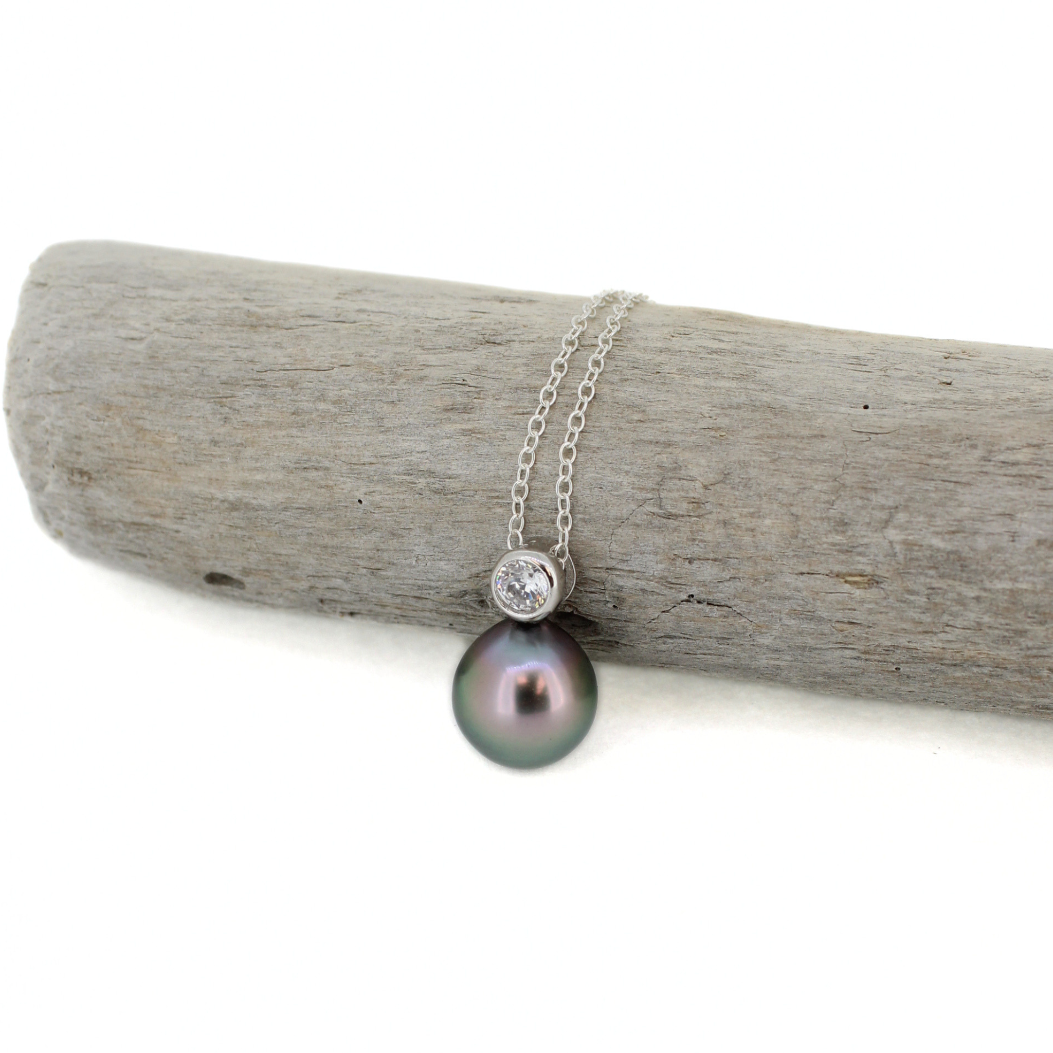 A silver necklace featuring a round dark gray pearl pendant and a bezel-set clear stone, displayed on a piece of weathered driftwood against a white background.