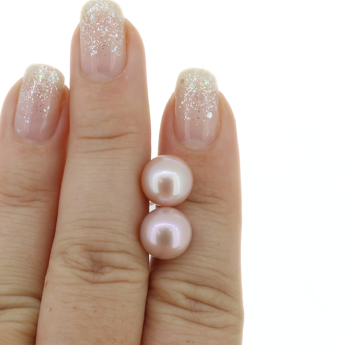 A close-up of a hand with glittery manicured nails holding a pair of pink Edison pearl stud earrings, on a white background.