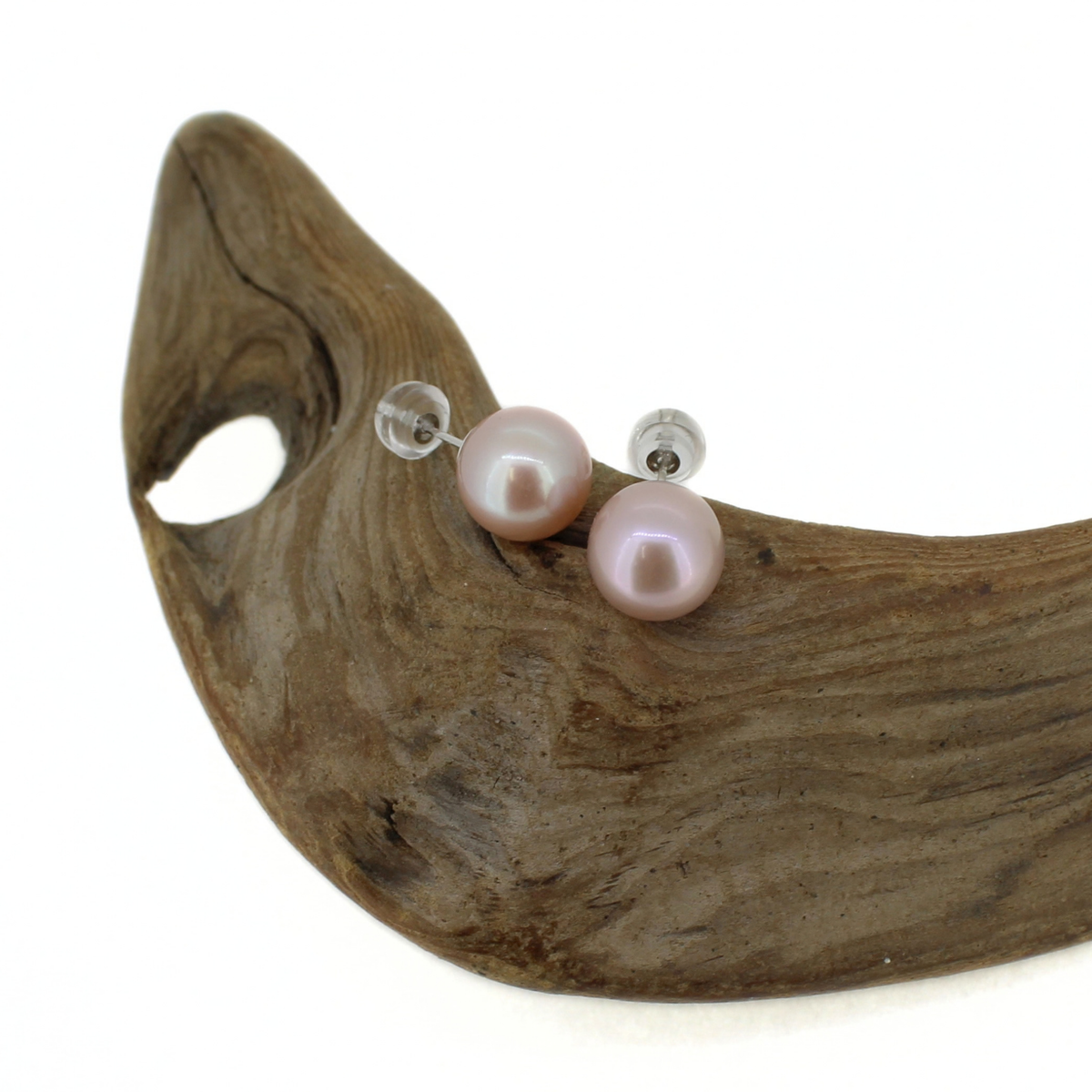 A pair of round light pink freshwater pearl stud earrings, resting on a curved piece of natural driftwood against a white background.