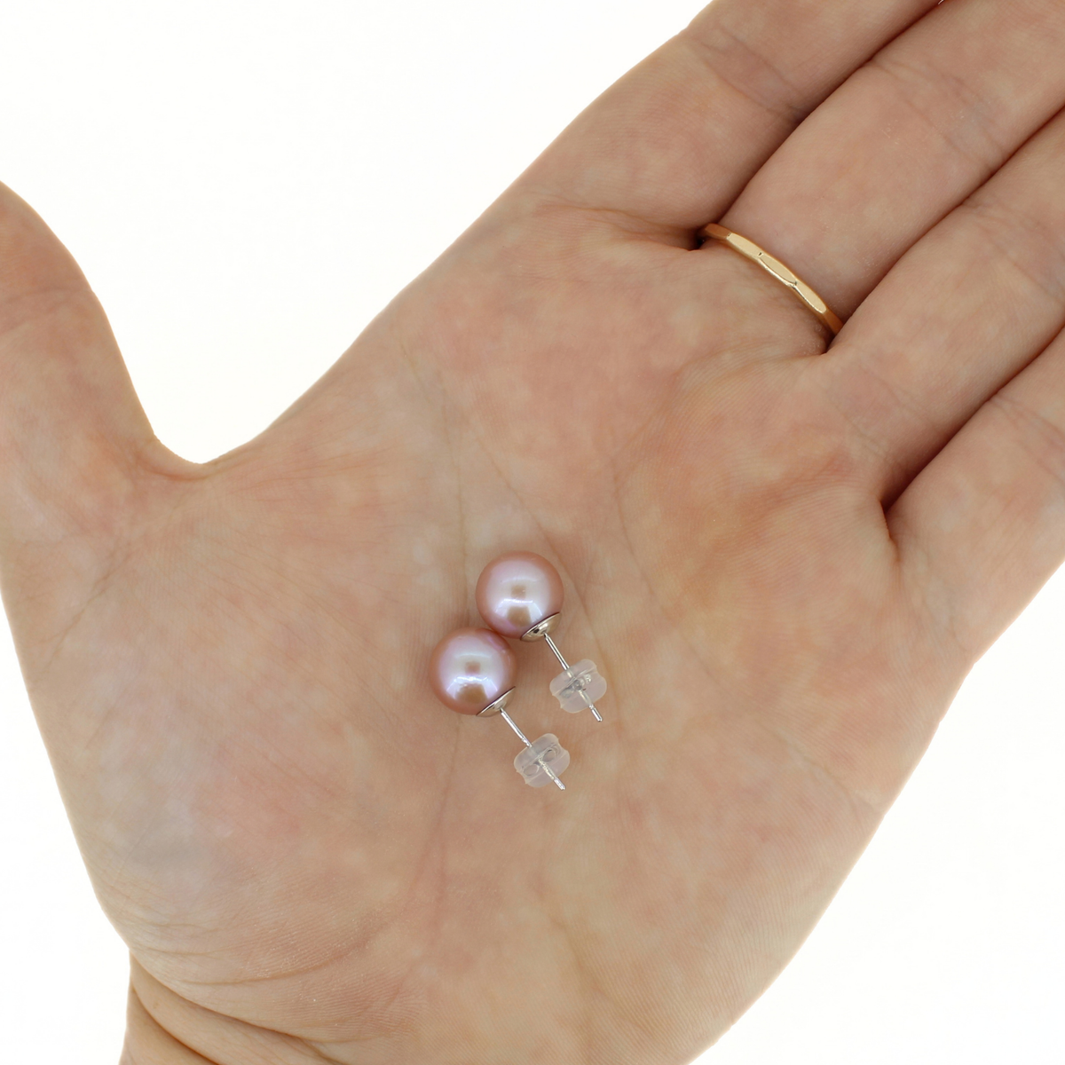 A pair of small pearl stud earrings with silver posts and clear square backings resting on an open hand. The pearls are a soft pink luster, and the hand also wears a thin gold ring on the finger.