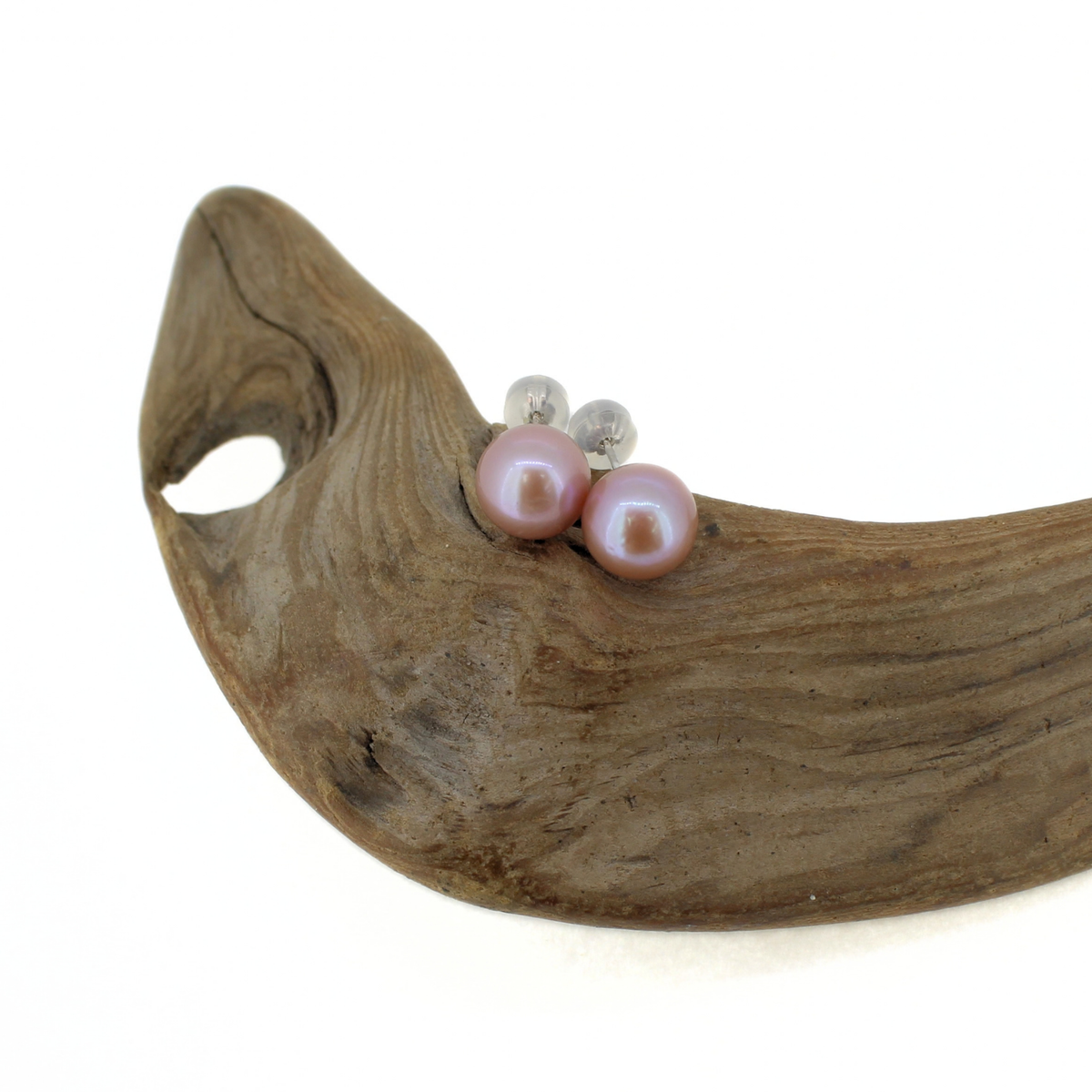 A pair of stud earrings with pink freshwater pearls on silver posts, displayed on a curved piece of driftwood against a white background.