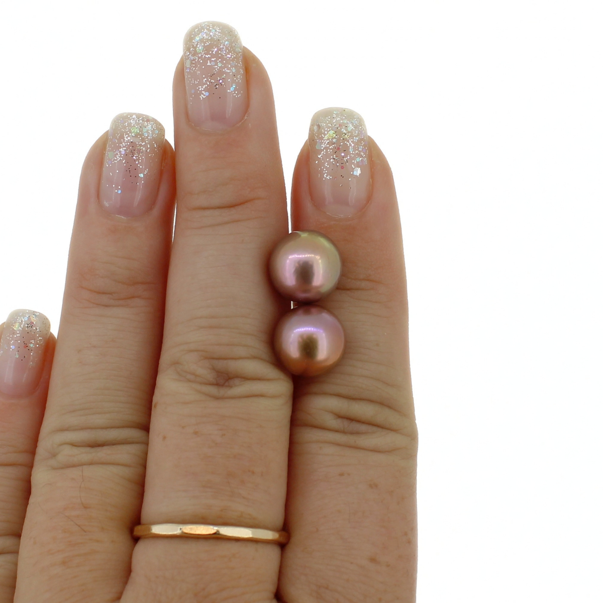 A close-up of a hand holding a pair of fresh water pearl stud earrings in a soft purple tone with metallic rose-gold reflections, alongside a thin gold band on the middle finger.