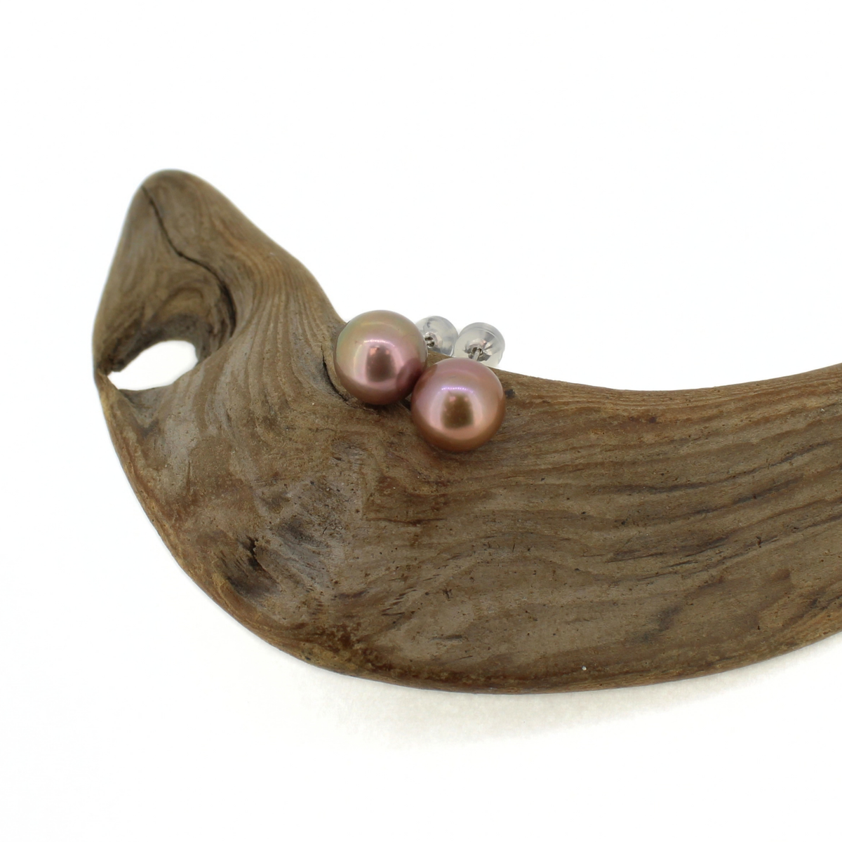 A pair of stud earrings with iridescent pinkish-purple freshwater pearls and small silver posts, displayed on a curved piece of driftwood against a white background.