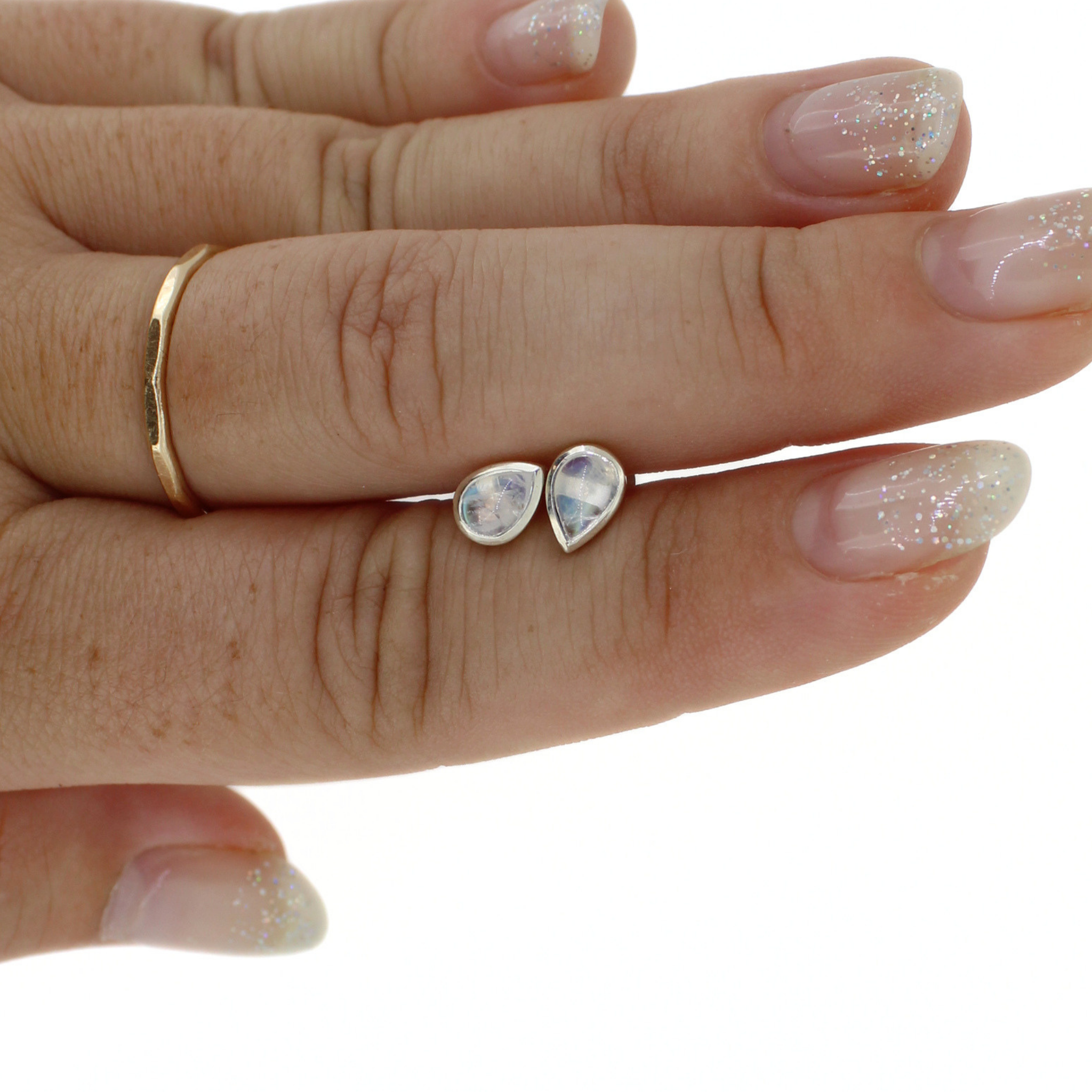A close-up of a hand holding a pair of small teardrop-shaped iridescent rainbow moonstone stud earrings against a plain white background.