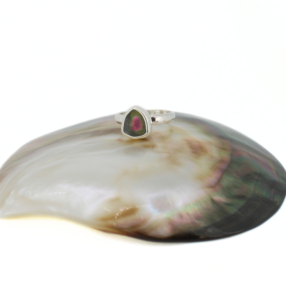 A silver ring with a triangular green and pink watermelon tourmaline stone, displayed on a large iridescent shell against a white background.