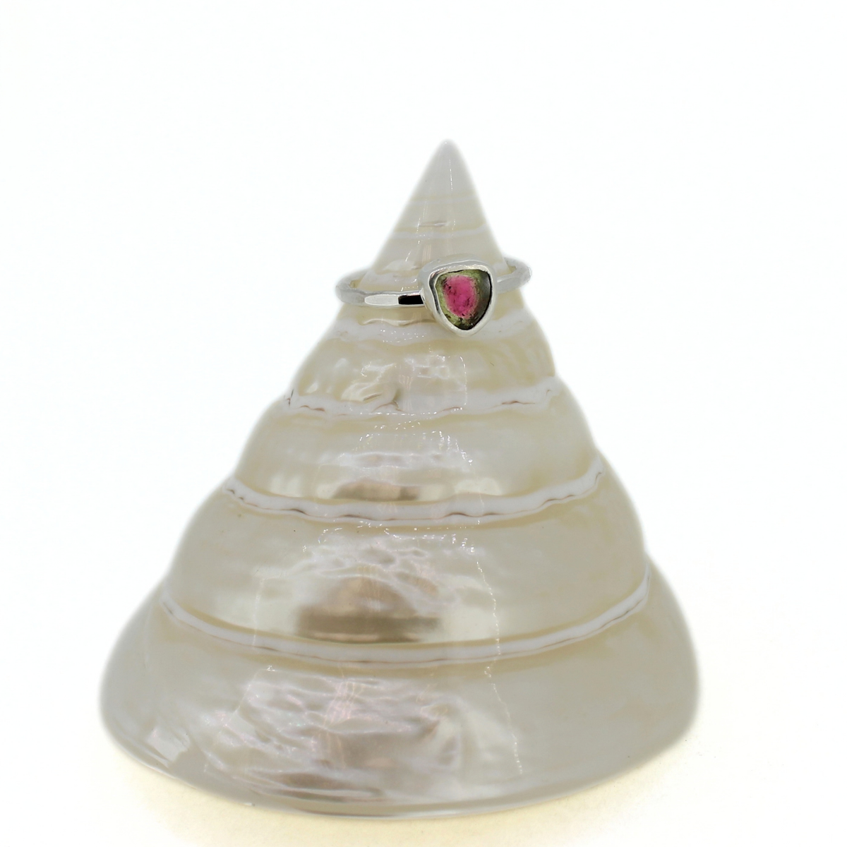 A silver ring with a small heart-shaped pink gemstone, displayed on a clear, spiral seashell stand against a white background.
