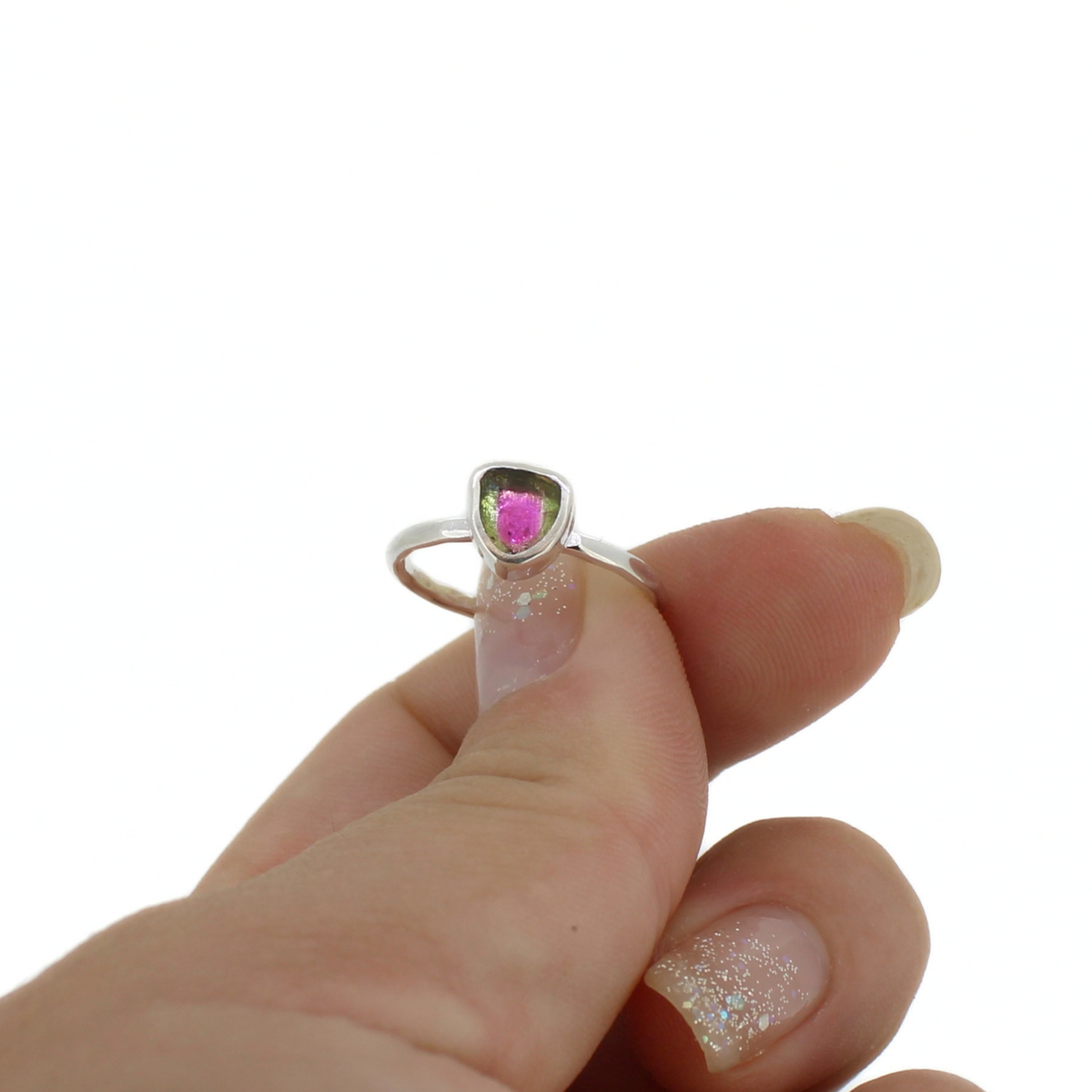 A hand wearing a silver ring with a small triangular bezel holding a bright pink and green gemstone, shown against a plain white background.