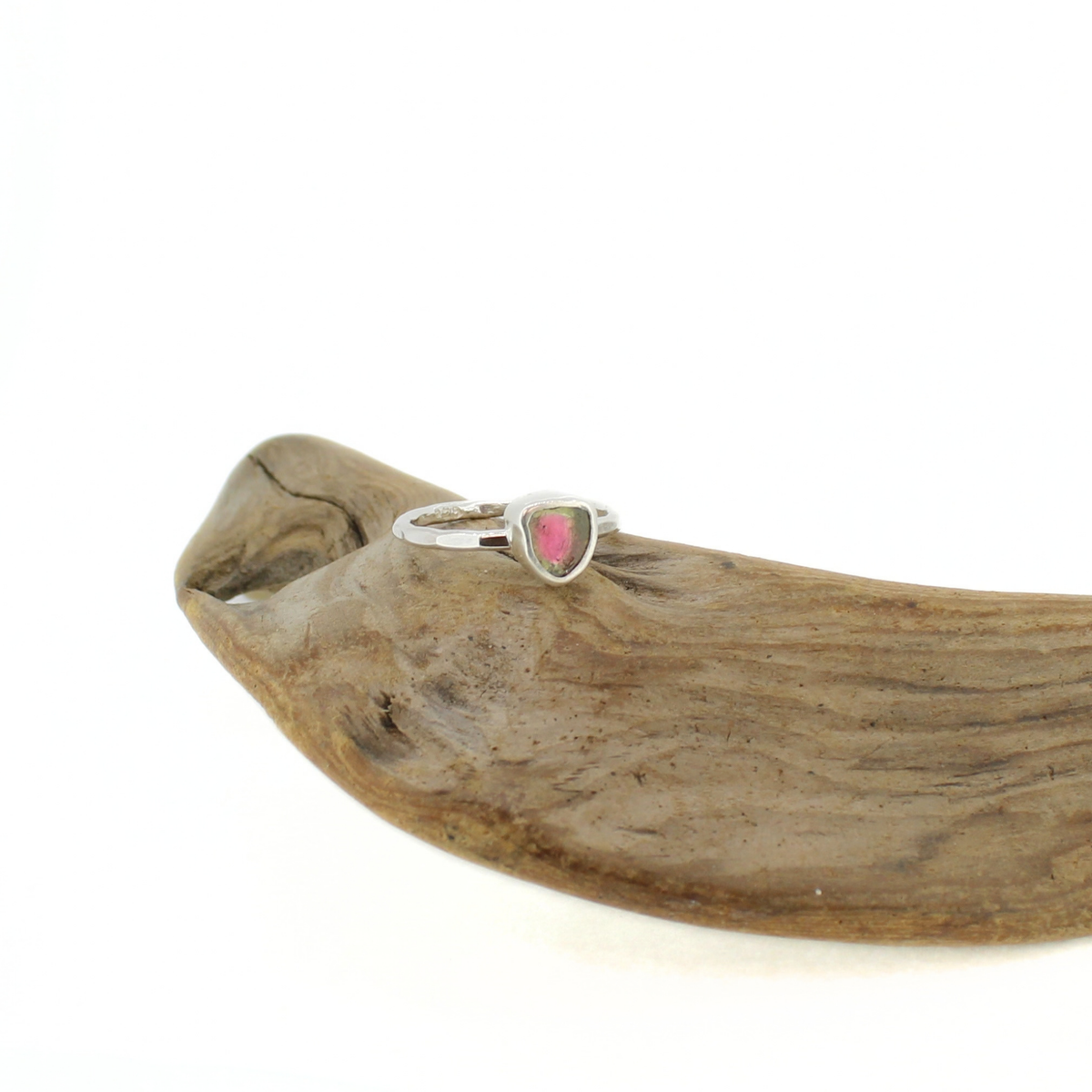 A silver ring featuring a small, triangular-shaped pink and green gemstone, displayed on a piece of natural driftwood against a white background.