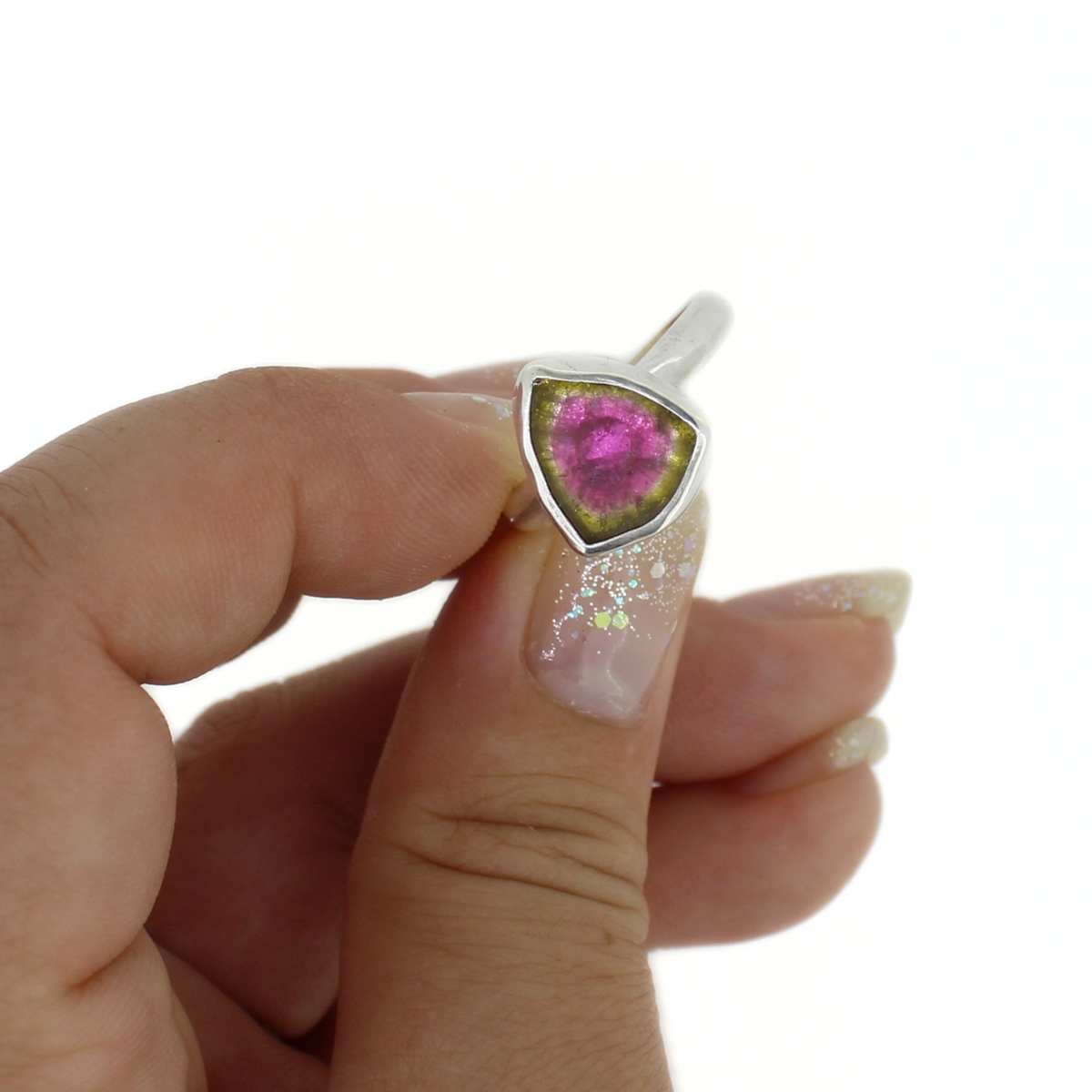 A hand with glittery nail polish holds a silver triangle-shaped ring featuring a vibrant pink gemstone with a green halo, set against a plain white background.