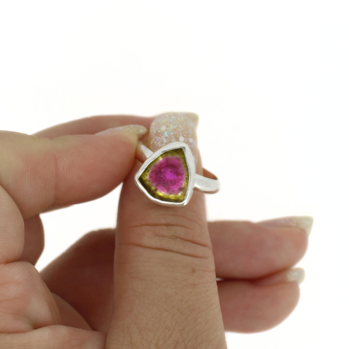 A hand wearing a silver ring with a triangular bezel set with a vibrant pink gemstone and an olive-green outer halo, shown against a plain white background.