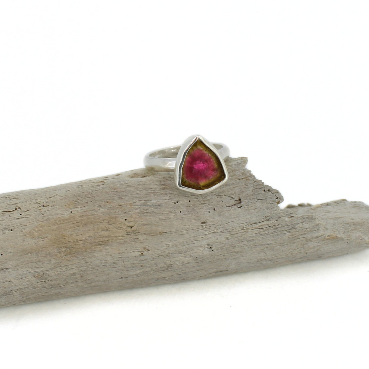 A silver ring with a triangular pink and green gemstone, resting on a piece of weathered driftwood against a white background.