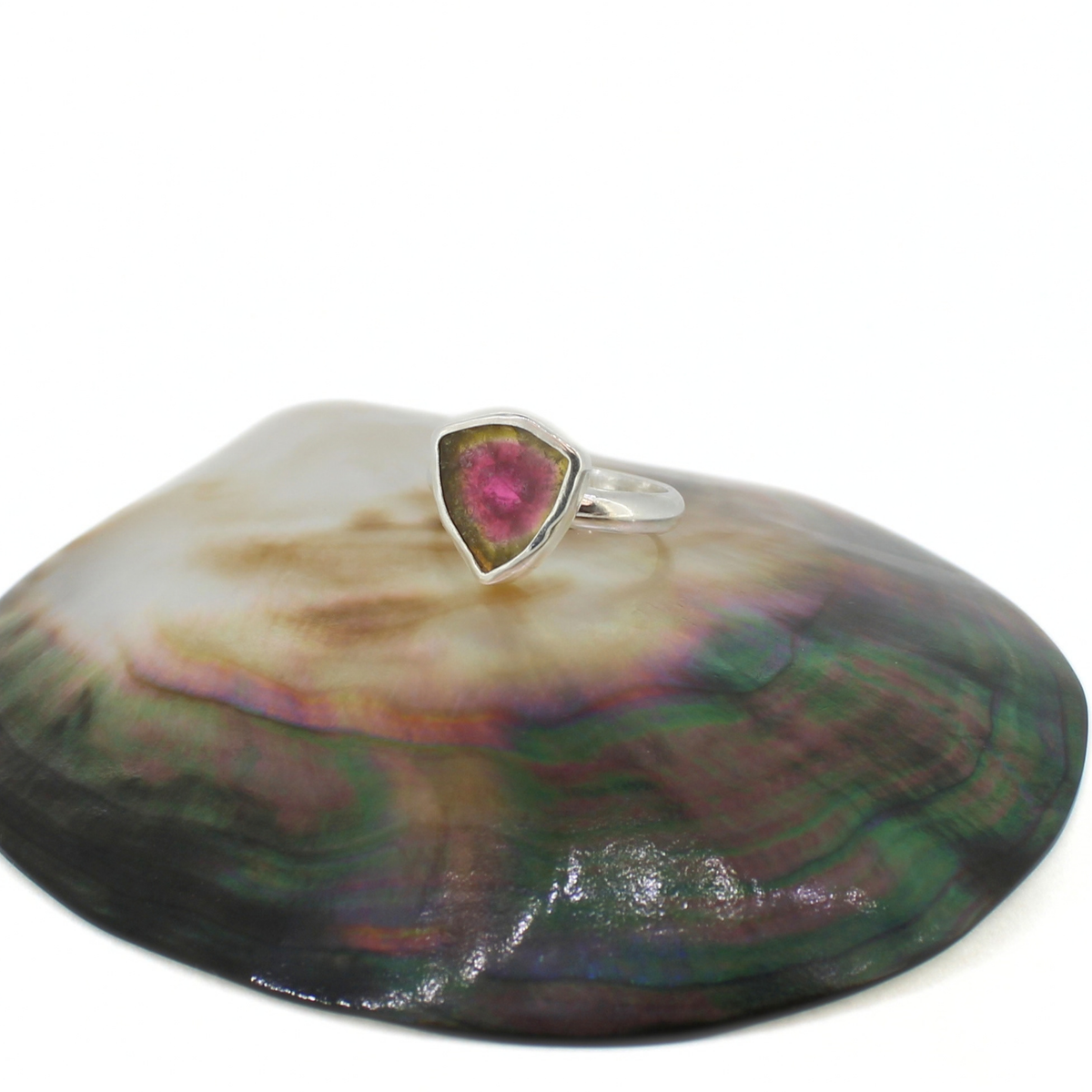 A silver ring with a triangular pink and green gemstone, displayed on top of a large iridescent abalone shell against a white background.