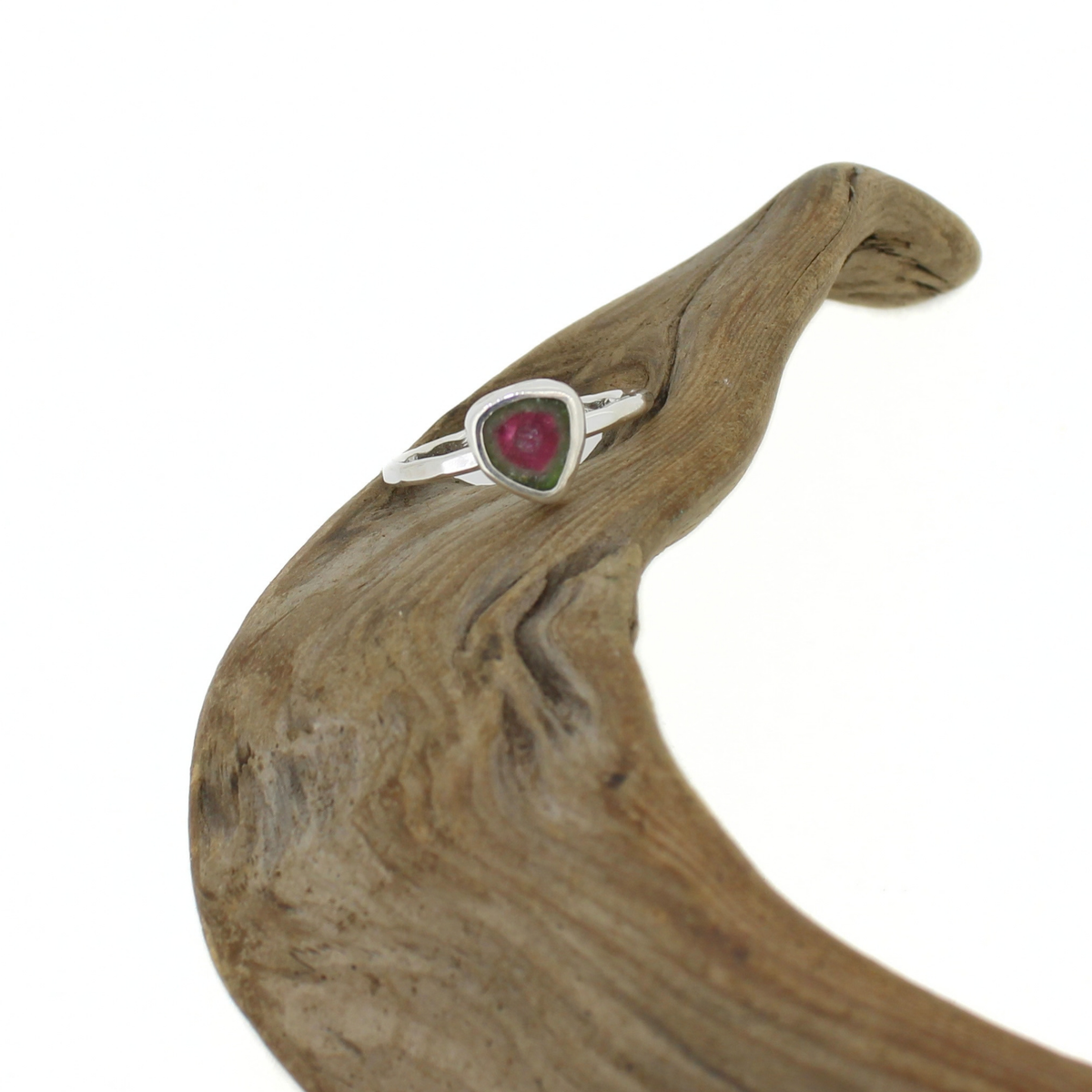 A silver ring with a triangular-shaped pink and green tourmaline gemstone, displayed on a curved piece of natural driftwood against a white background.