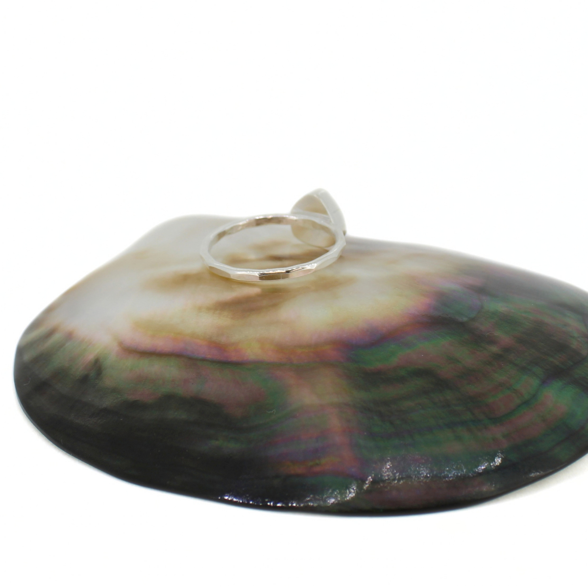 A polished silver ring with a triangular bezel-set stone, displayed on top of a large iridescent abalone shell dish against a white background.