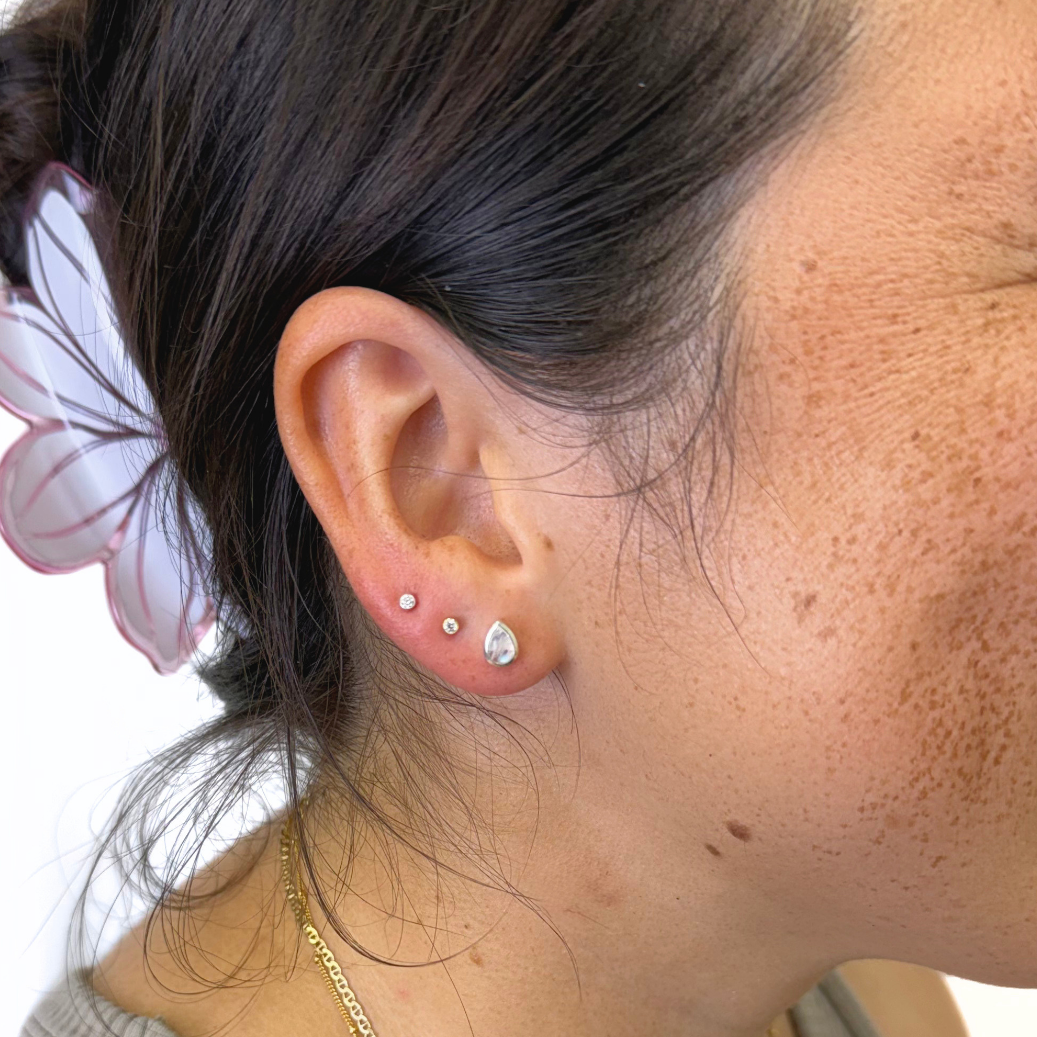 A close-up of a person's ear showcasing three silver stud earrings of varying sizes, including a teardrop-shaped stud. The person has dark hair tied back with a white and pink hair clip and is wearing a delicate gold chain necklace.
