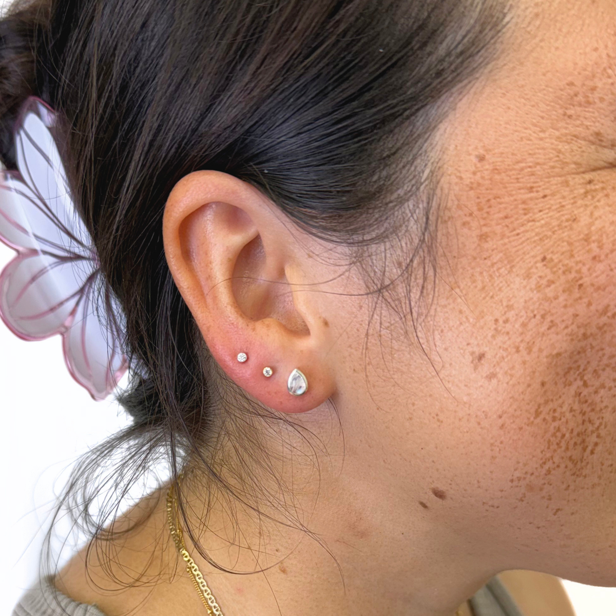 A close-up of a person's ear showcasing three silver stud earrings of varying sizes, including a teardrop-shaped stud. The person has dark hair tied back with a white and pink hair clip and is wearing a delicate gold chain necklace.